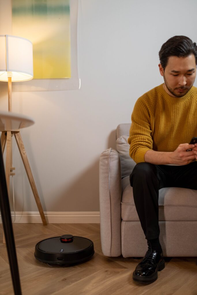 pexels-photo-6856816-6856816 A man in a yellow sweater uses his phone on a sofa with a robot vacuum nearby.